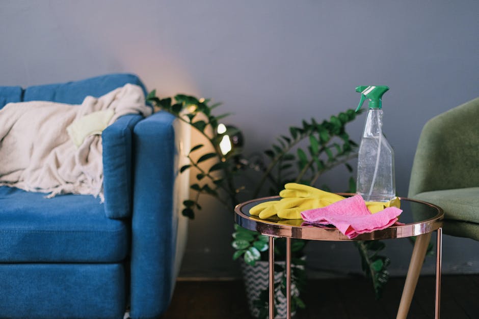 An indoor scene featuring a blue upholstered sofa with a beige blanket draped over its armrest, positioned next to a small round side table with a rose gold finish. The table holds cleaning supplies i